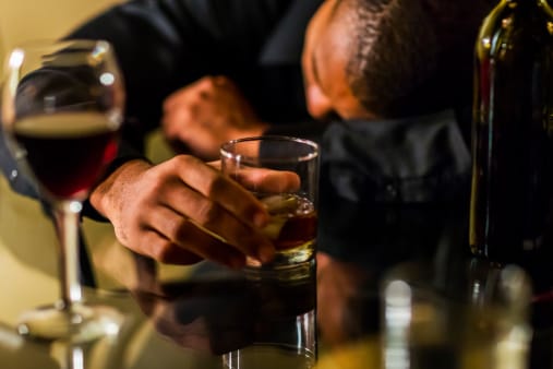 Guy laying on a table passed out with drink in hand, looks like he might need alcohol rehab